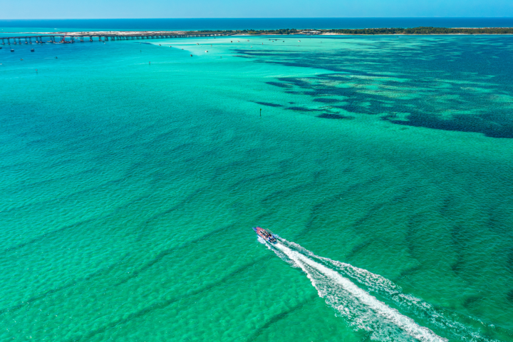 Aerial view of a boat traveling on clear turquoise water near a distant shoreline.
