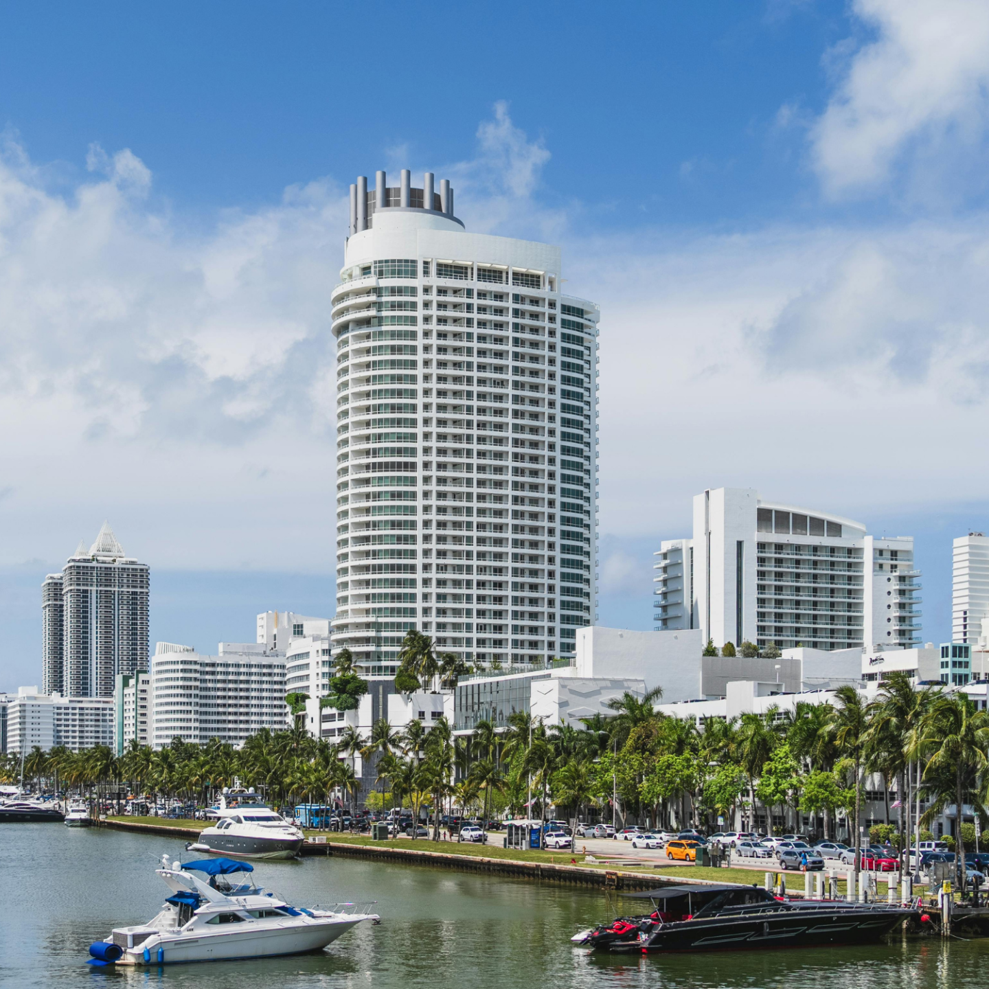 Modern cityscape with high-rise buildings, palm trees, and boats on a sunny day.
