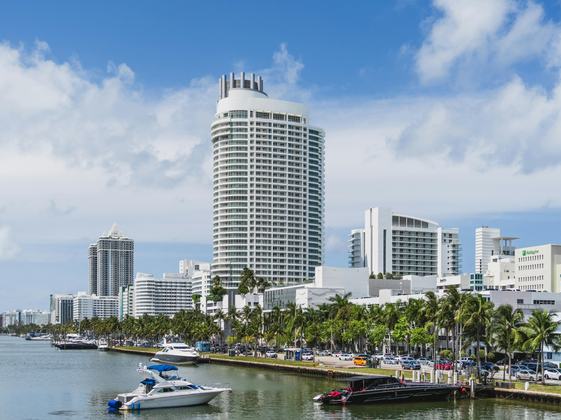 Modern cityscape with high-rise buildings, palm trees, and boats on a sunny day.