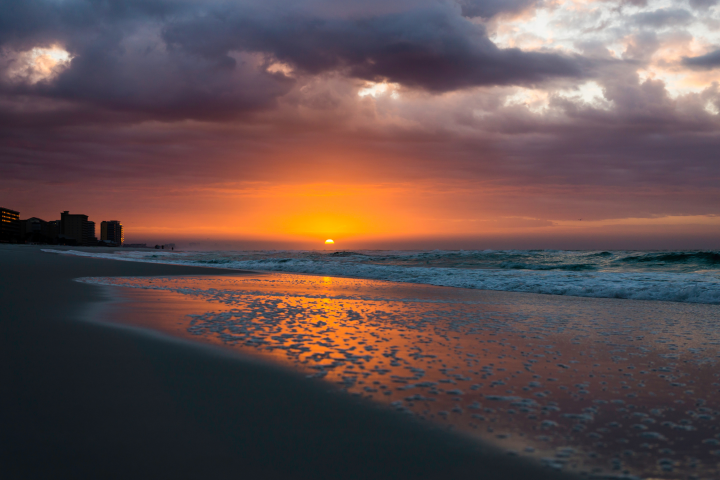 Sunset over ocean with buildings silhouetted, waves and foam on sandy beach.