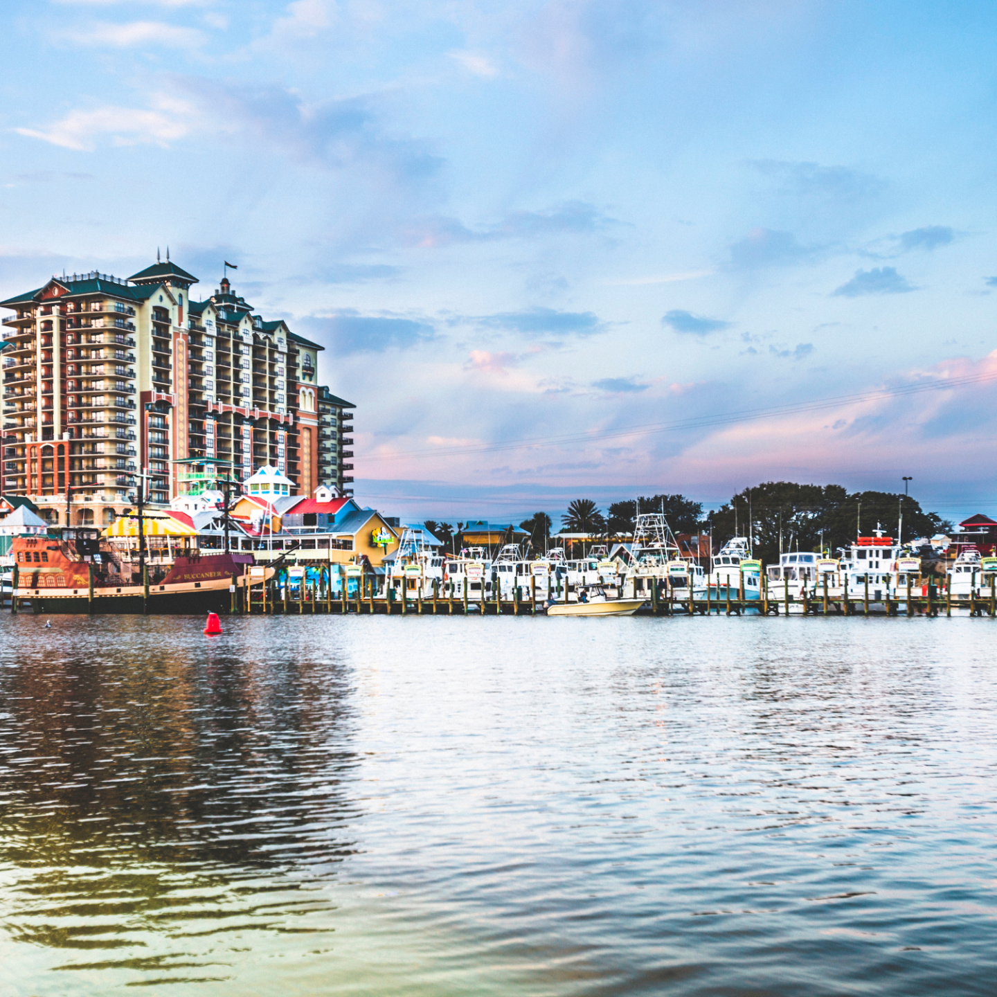 Waterfront scene with tall buildings, colorful shops, boats, and a lookout tower under a blue sky.