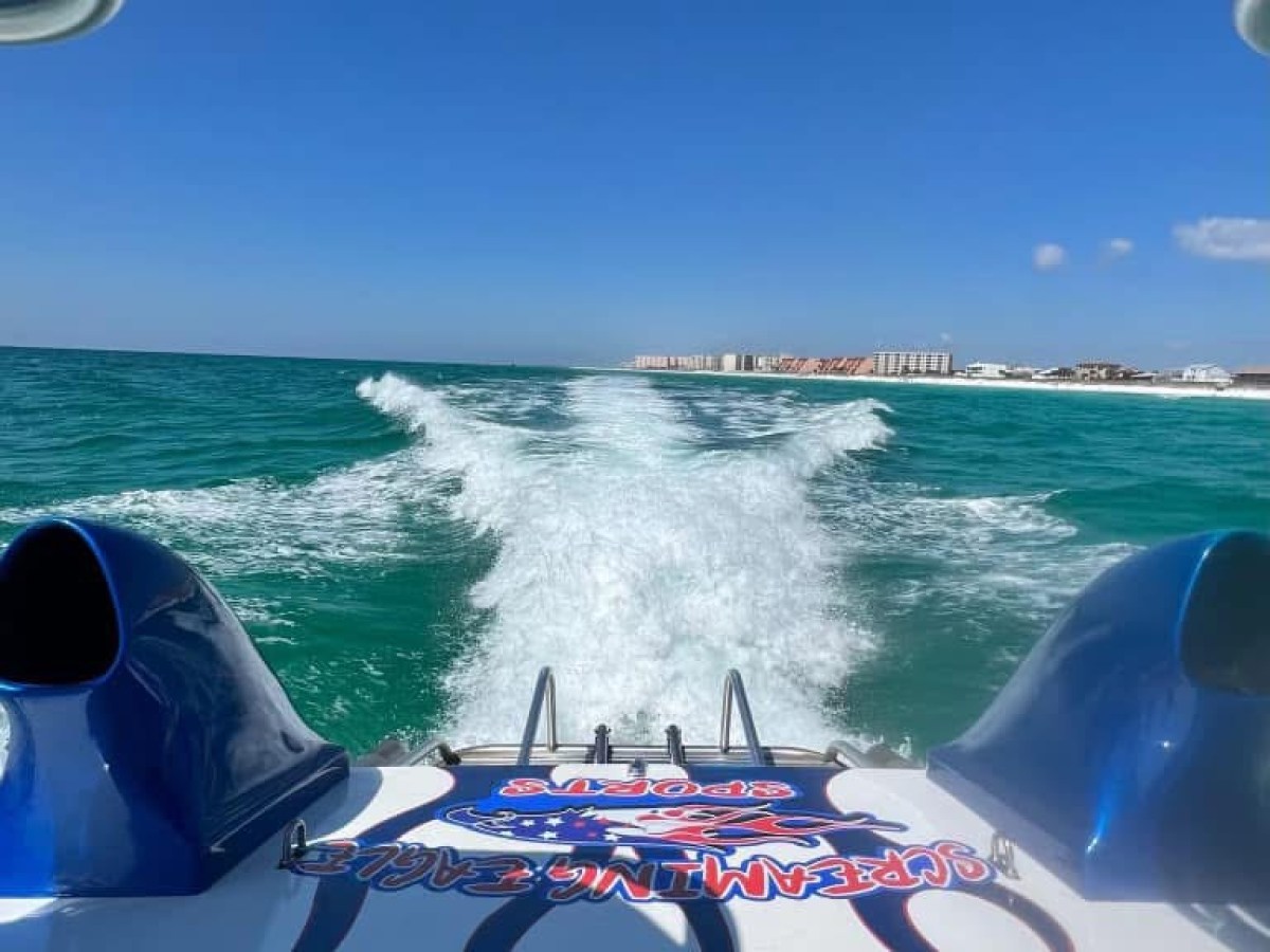 Back of a speedboat on a sunny sea with wake trails and distant buildings.