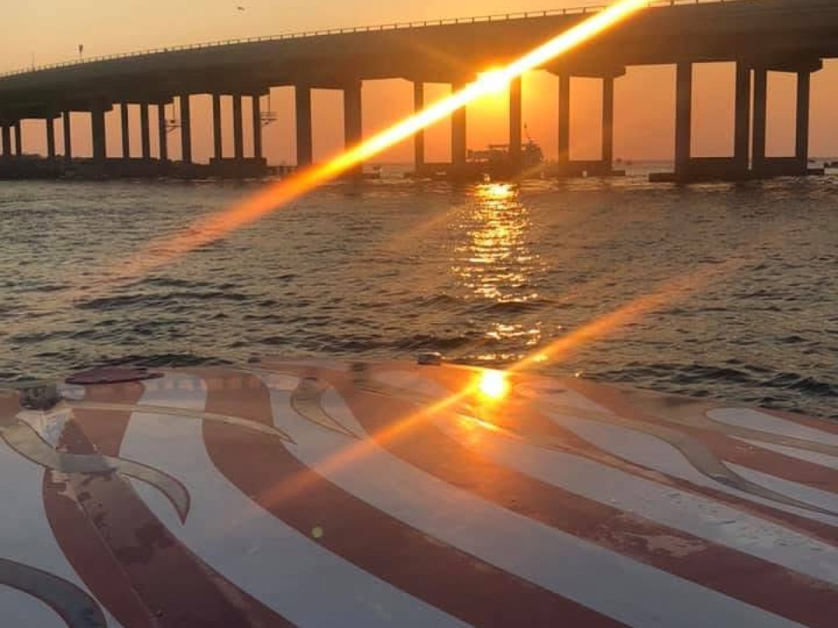 Sunset over a bridge with a striped boat in the foreground on the water.