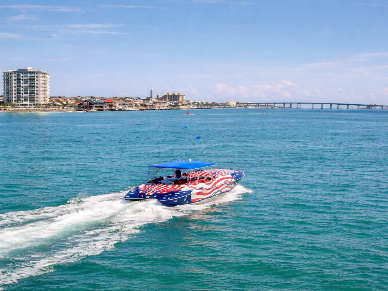 Boat with American flag design moves on blue water near coastal buildings and a bridge.
